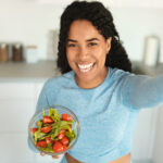 Healthy eating concept. Happy sporty black lady holding bowl with fresh vegetable salad and taking selfie in kitchen